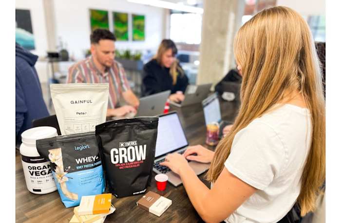Several people at a table testing various protein powders mixed with water and recording their notes on laptops